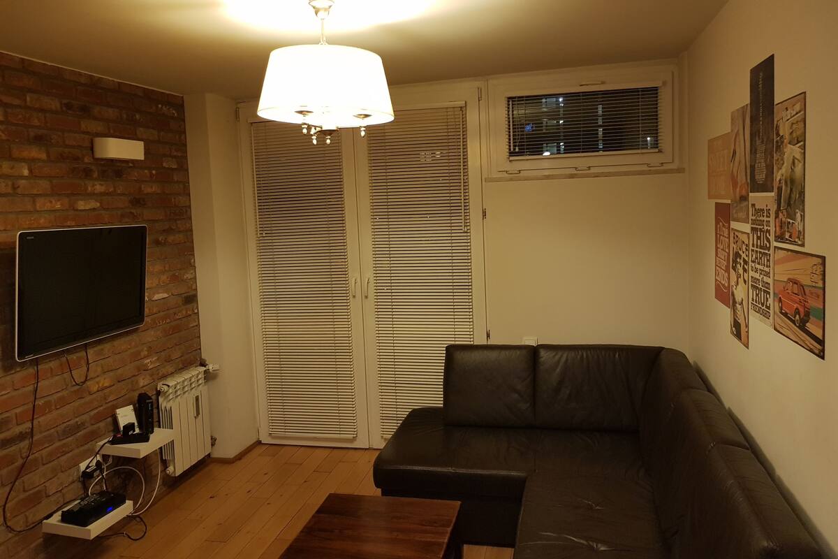 A living room with a black leather sofa, a wooden coffee table, a flat-screen TV mounted on a brick wall, a ceiling light, and a window with blinds.