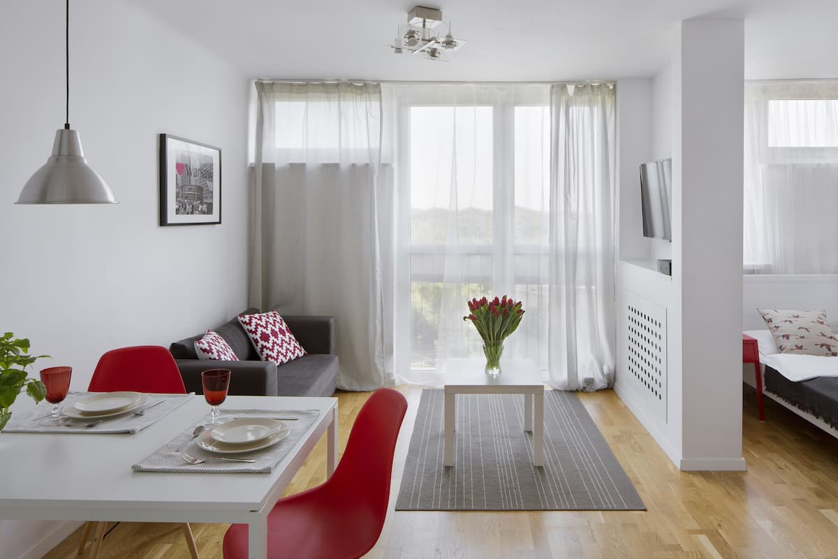 A modern living room with a dining area, featuring a sofa, red chairs, a dining table set with plates and glasses, and a vase with flowers.