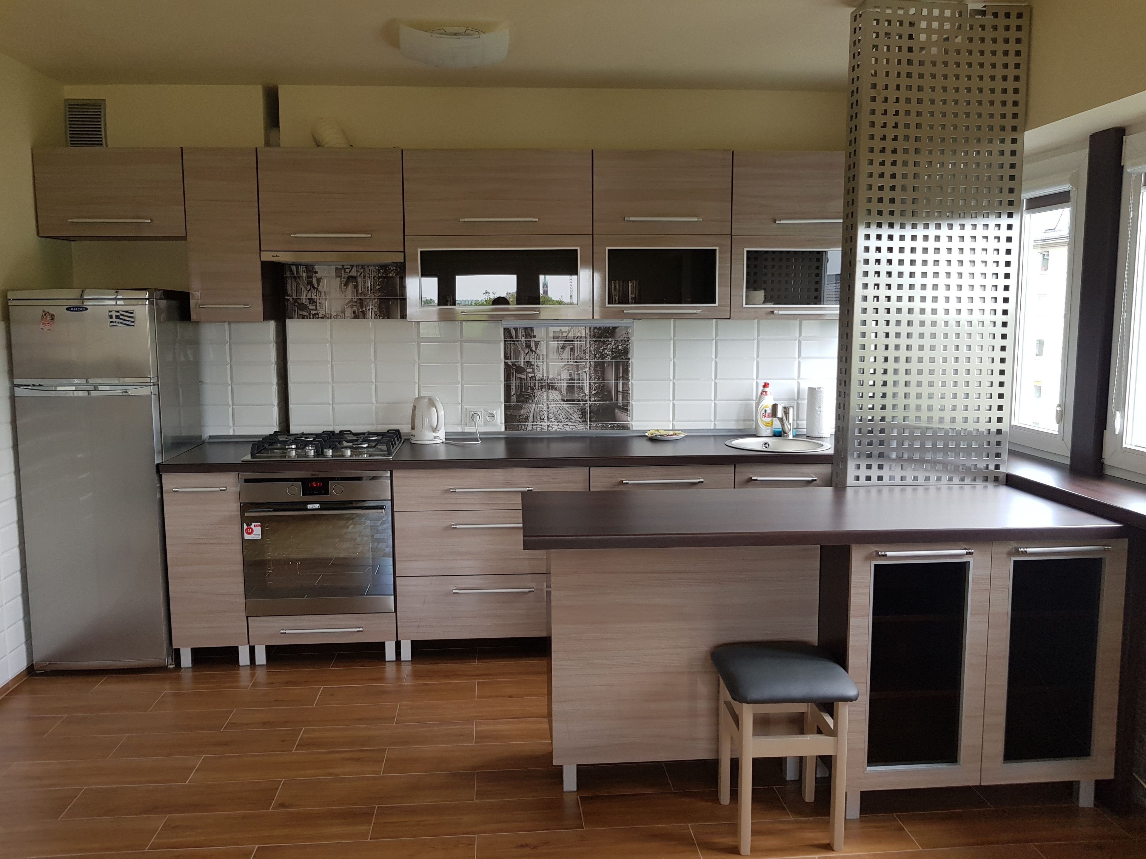 A modern kitchen with wooden cabinets, a stainless steel refrigerator, a built-in oven, a gas stove, and a sink. There is a window with a view of the outside.