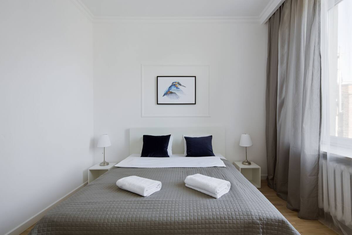A neatly made bed with a grey quilt, two pillows, and two rolled towels. There is a framed picture of a bird on the wall above the bed.