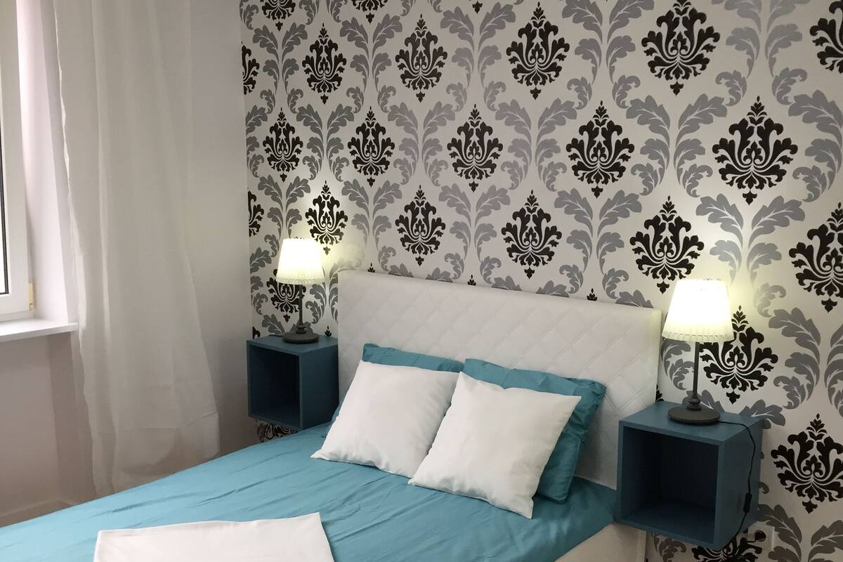 A bed with a blue and white bedspread, two white pillows, and a white throw blanket. There are two bedside tables with lamps, and a white furry rug on the floor.