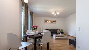 Un coin repas avec une table dressée pour quatre personnes, des chaises blanches, un vase avec des fleurs et, au fond, un canapé moderne.