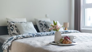 Un lit bien fait, recouvert d’une couvre-lit à motifs, un vase rempli de fleurs et une assiette de pâtisseries, le tout posé sur une table de chevet, à côté d’une tasse.