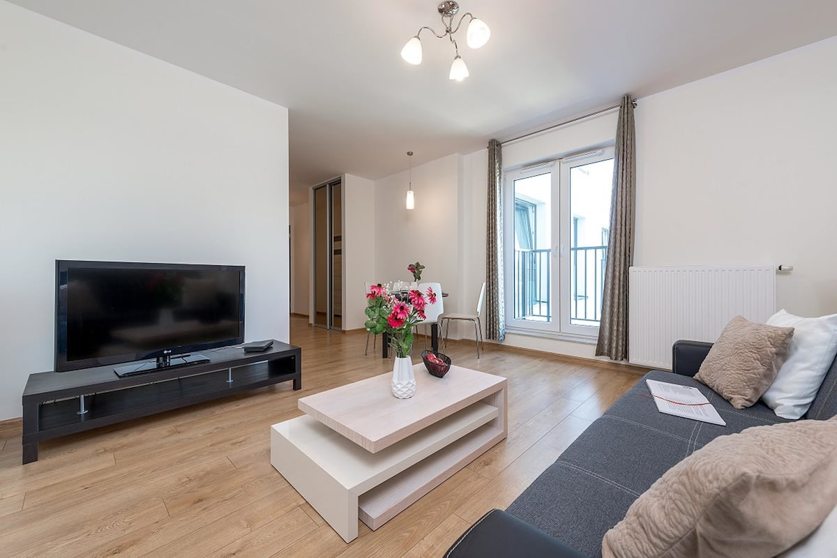 Een moderne woonkamer met een flatscreen televisie, een houten salontafel met een bloevas, een bank en een groot raam met gordijnen.