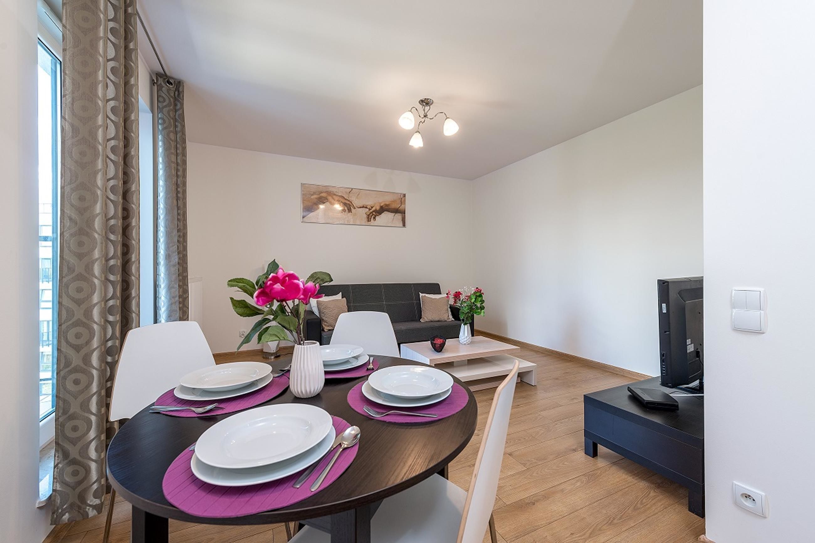 A modern dining area with a round table set for four, a sofa, and a TV in the background.
