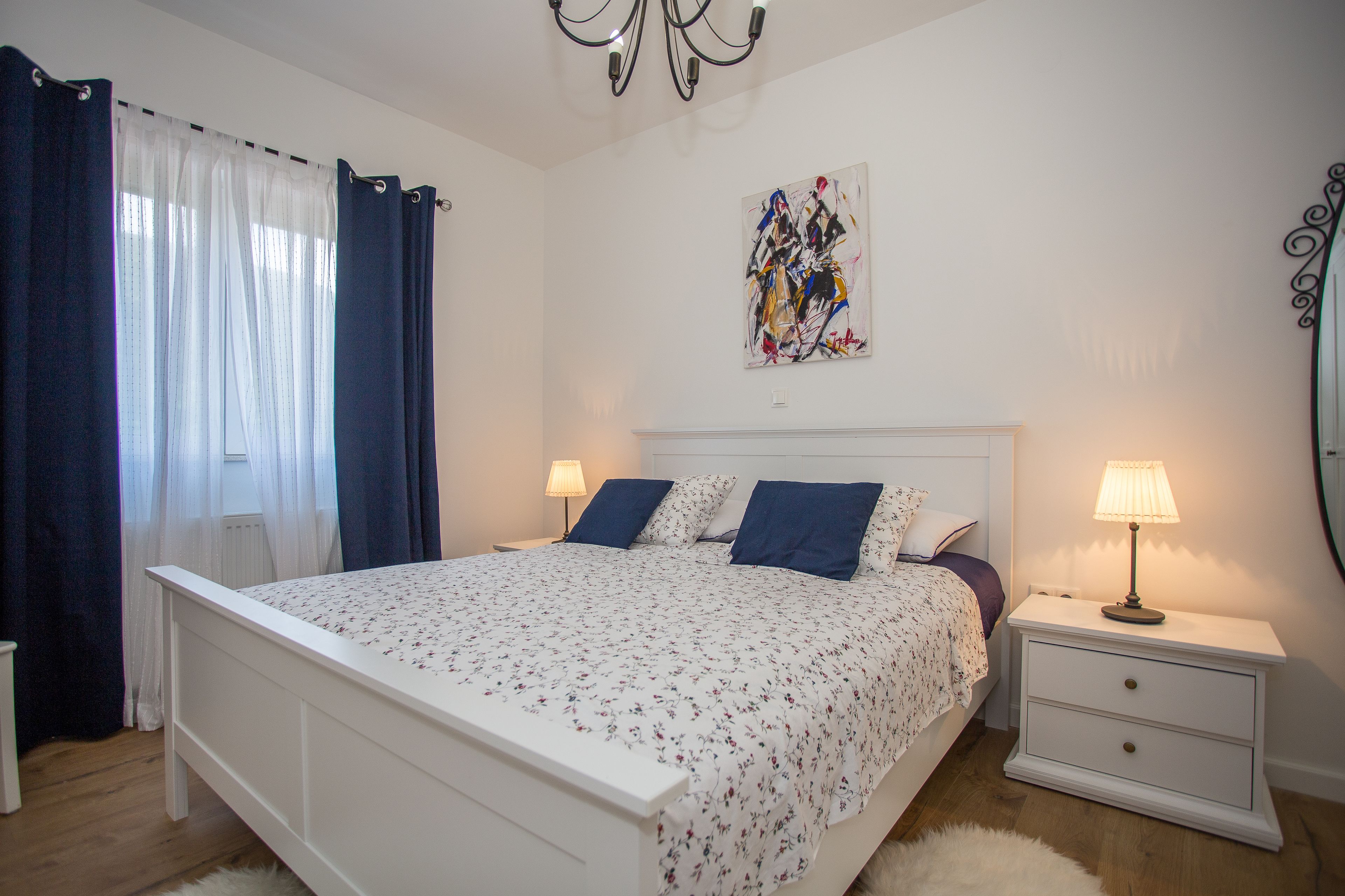 Een slaapkamer met een wit bed, blauwe gordijnen, een nachtkastje met een lamp en een kunstwerk aan de muur.
