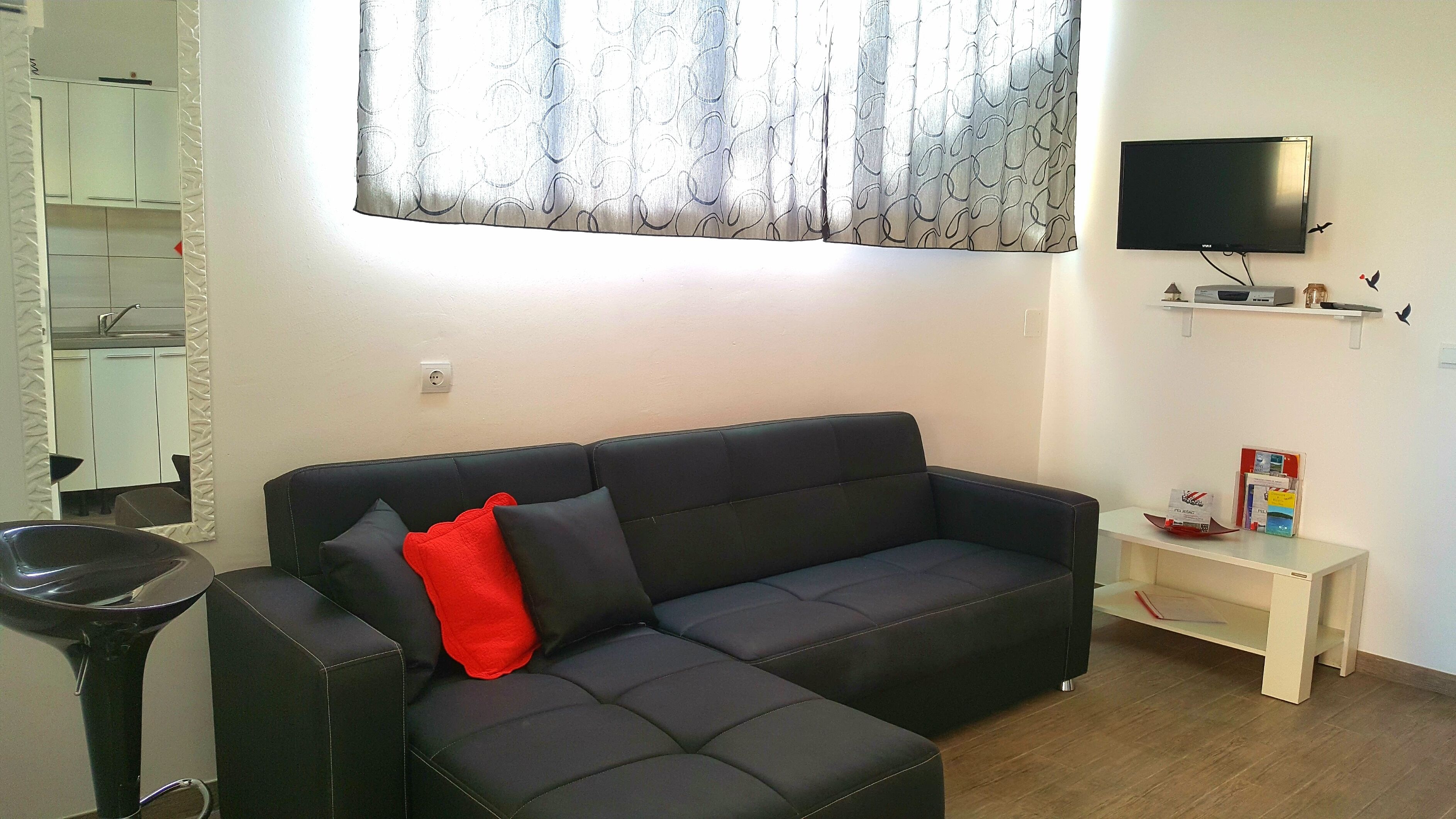 A modern living room with a black leather sofa, a small white side table, a flat-screen TV mounted on the wall, and a window with patterned curtains.