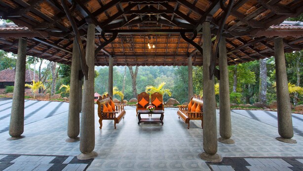A covered outdoor seating area with stone pillars, wooden furniture, and a view of a lush garden. - Spices Lap Thekkady (Udumbanchola)