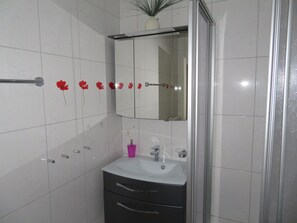 Un baño con paredes de azulejos blancos, mampara de ducha de vidrio, tocador negro con lavamanos blanco y espejo sobre el tocador.