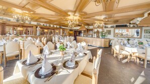 A spacious dining area with multiple tables set for a meal, wooden ceiling beams, and chandeliers. - Villa Rhenus (Ginsheim-Gustavsburg)
