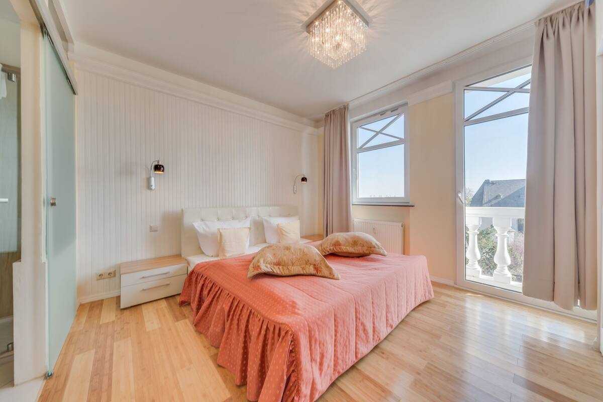 A bedroom with a large bed, a chandelier, a view of the outside, and a window with curtains.