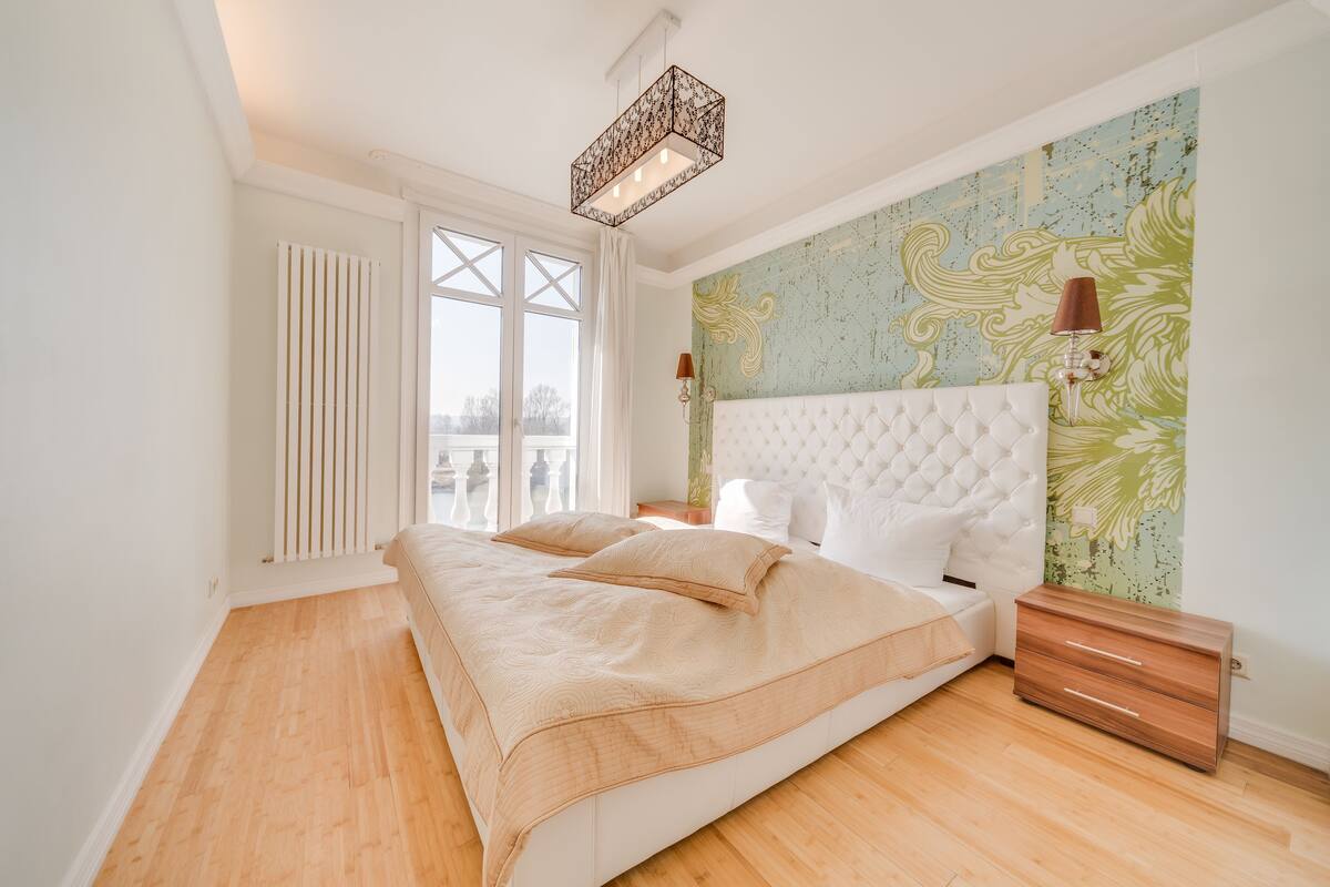 A bedroom with a large bed, a nightstand, a dresser, a radiator, and a window with curtains.