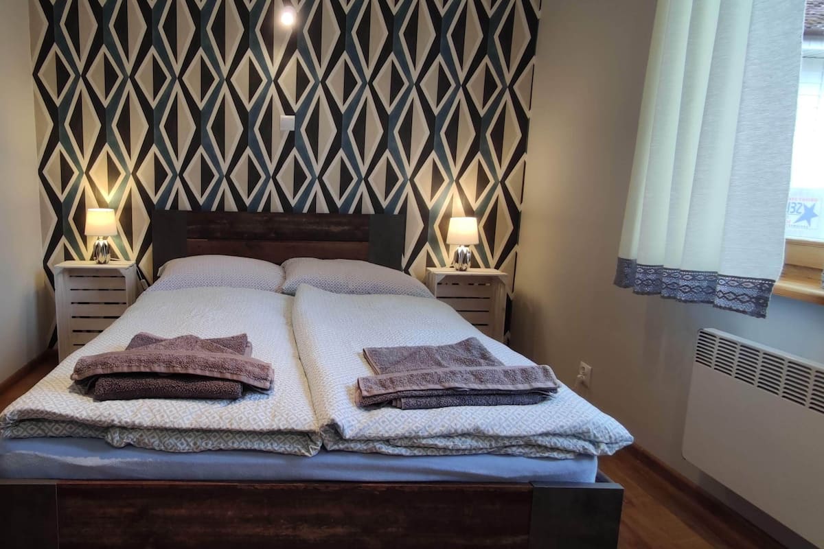 A double bed with two pillows and two folded towels, a wooden headboard, two bedside tables with lamps, a geometric patterned wall, and a window with curtains.