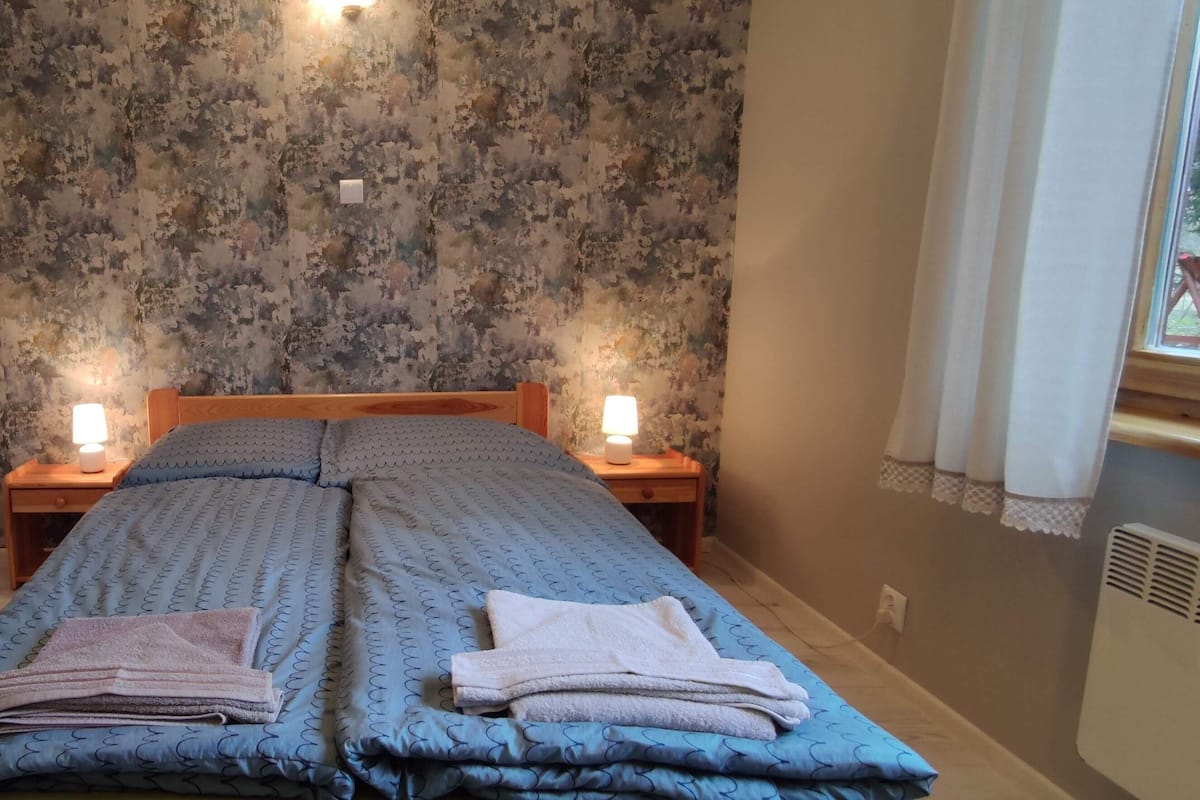 A room with a bed, two bedside tables with lamps, a floral-patterned wall, and a window with curtains.