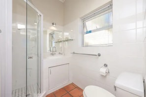 Ein Badezimmer mit Duschkabine aus Glas, Toilette in Weiß, kleinem Fenster mit Jalousien und Handtuchhalter.