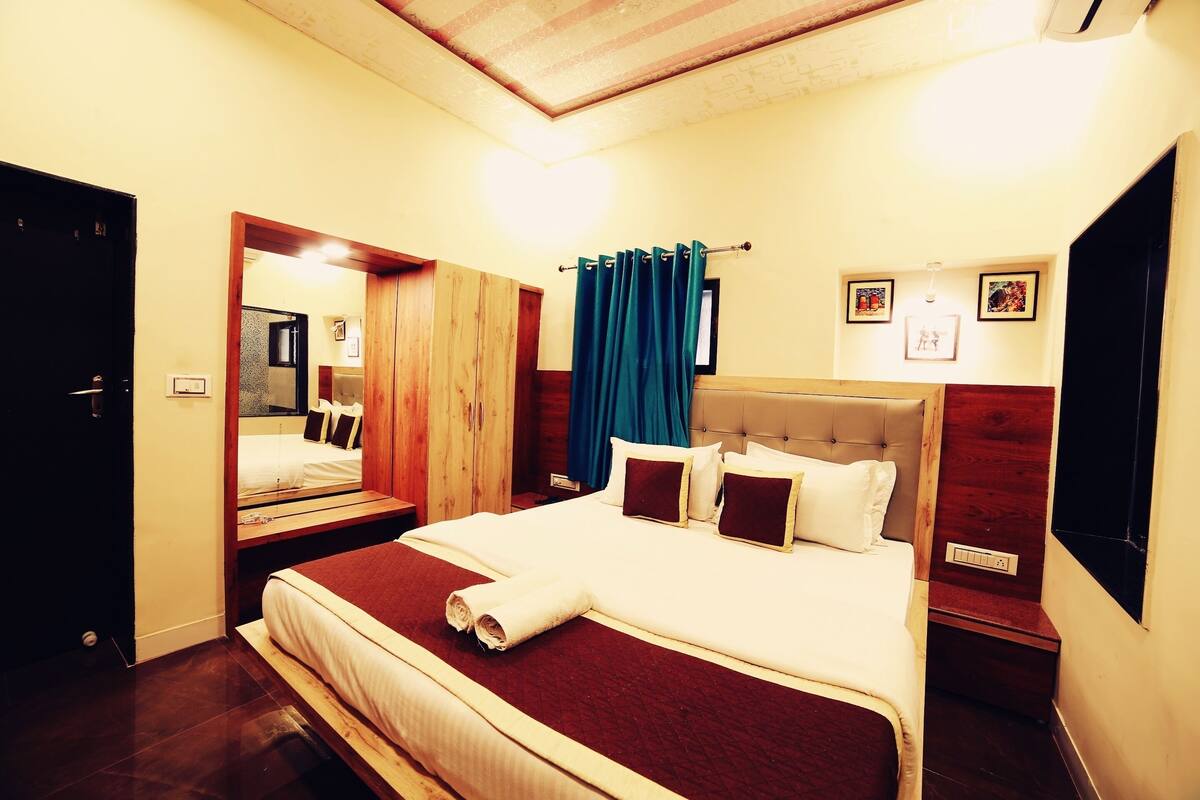 Une chambre d’hôtel avec un grand lit, une tête de lit en bois, un miroir, une fenêtre avec des rideaux et un téléviseur fixé au mur.