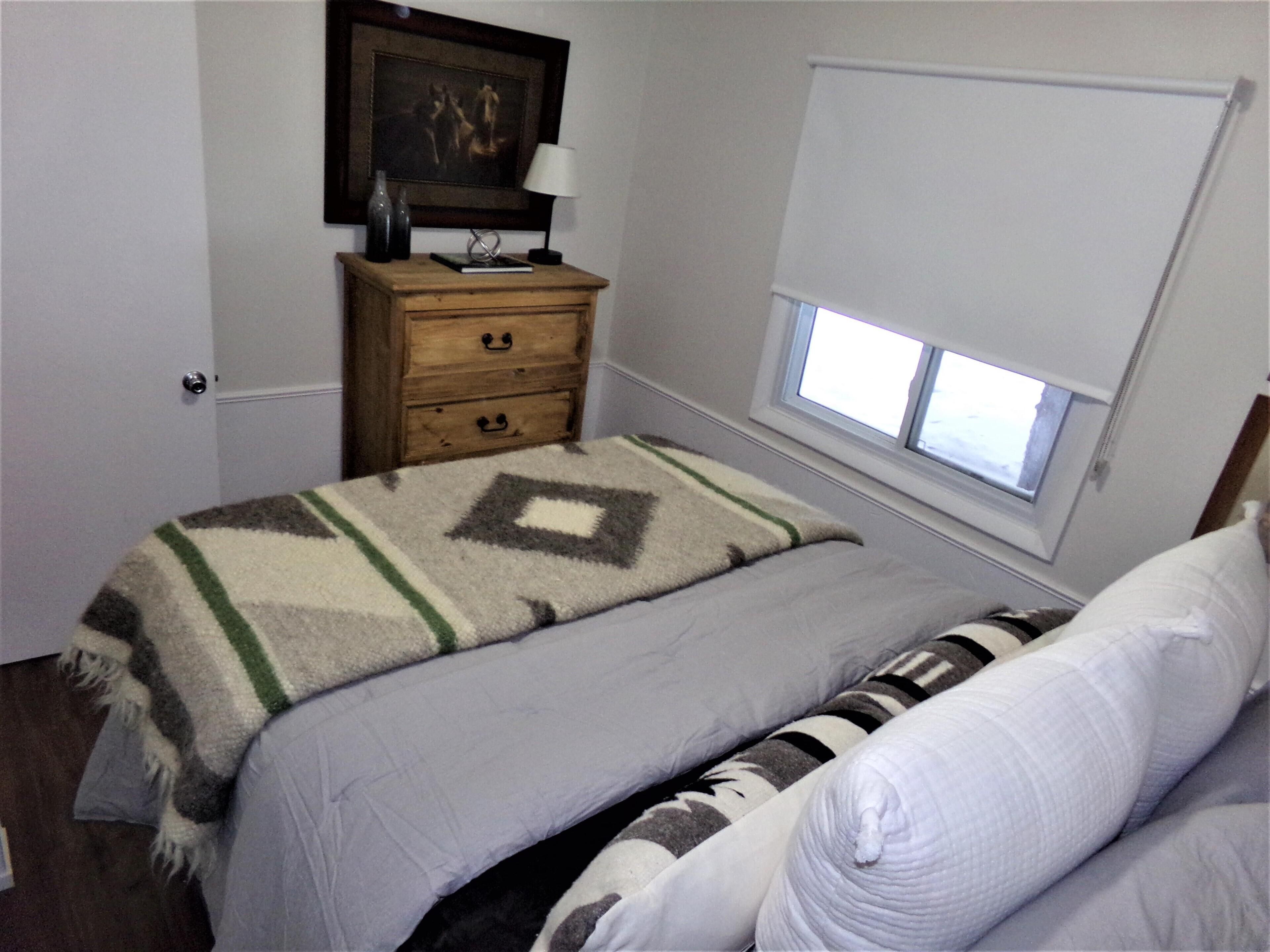 A bedroom with a bed, a nightstand, a window with blinds, a door, and a framed picture on the wall.