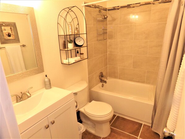 Un baño con inodoro, lavabo y bañera. Hay una cortina de ducha, un espejo y un estante con artículos de aseo.