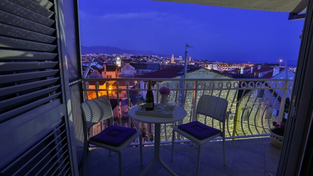 Un balcón con dos sillas y una mesa, con vista a un paisaje urbano al atardecer.