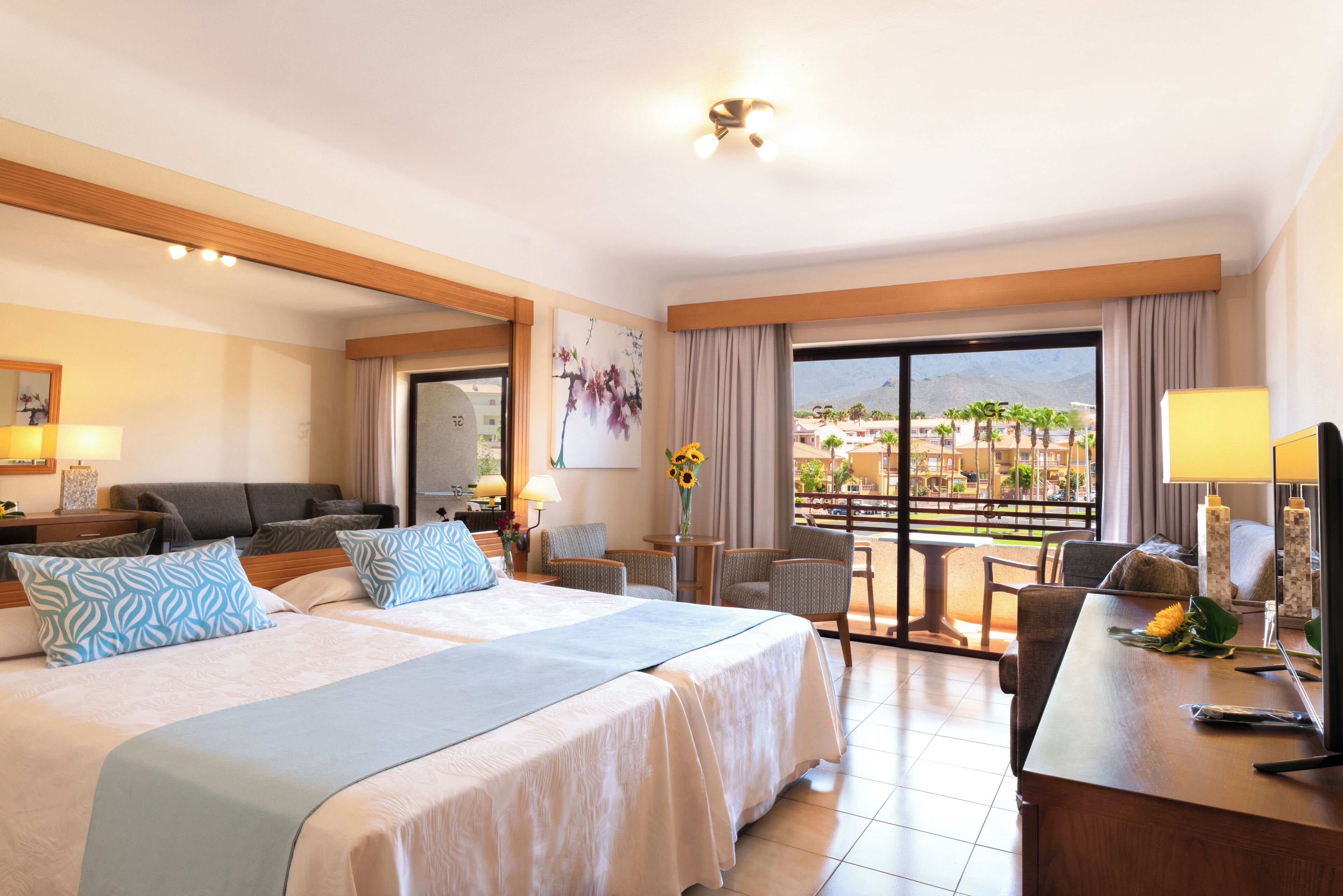 A hotel room with two beds, a desk, a chair, a sofa, a balcony with a view, and a dining area.