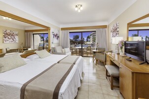 A hotel room with two beds, a desk with a TV, a balcony with a pool view, and artwork on the walls.