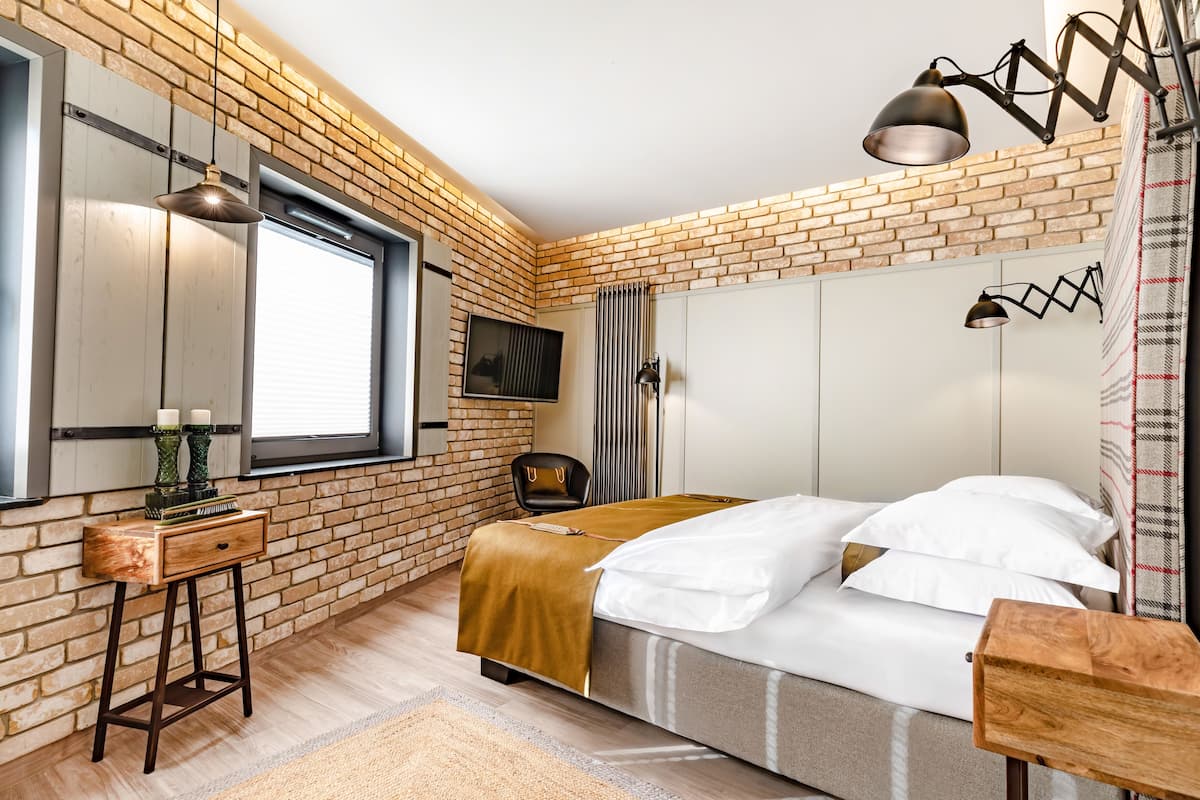 Un dormitorio con pared de ladrillo, una cama con ropa de cama blanca, una mesita de noche de madera, un escritorio y una lámpara colgante.
