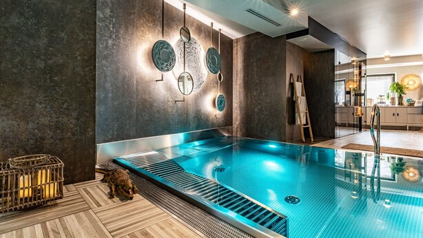Piscina interior moderna con superficie de agua cristalina, suelo de madera y pared de piedra con espejos circulares.