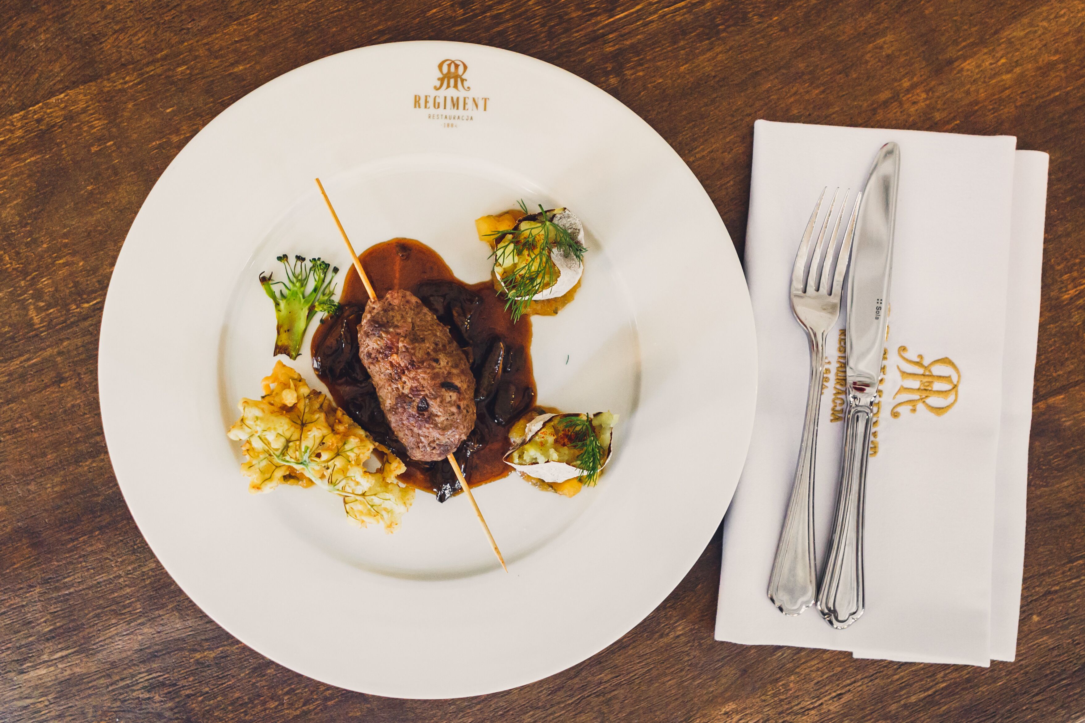Ein weißer Teller mit einem Steak, Gemüse, Soße und einem gefalteten Servietten mit Besteck auf einem Holztisch.