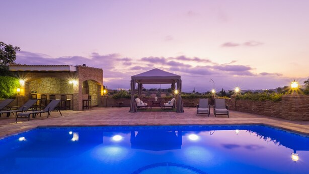 Una piscina con lettini e un gazebo, circondata da mura in pietra e un edificio in pietra con illuminazione esterna.