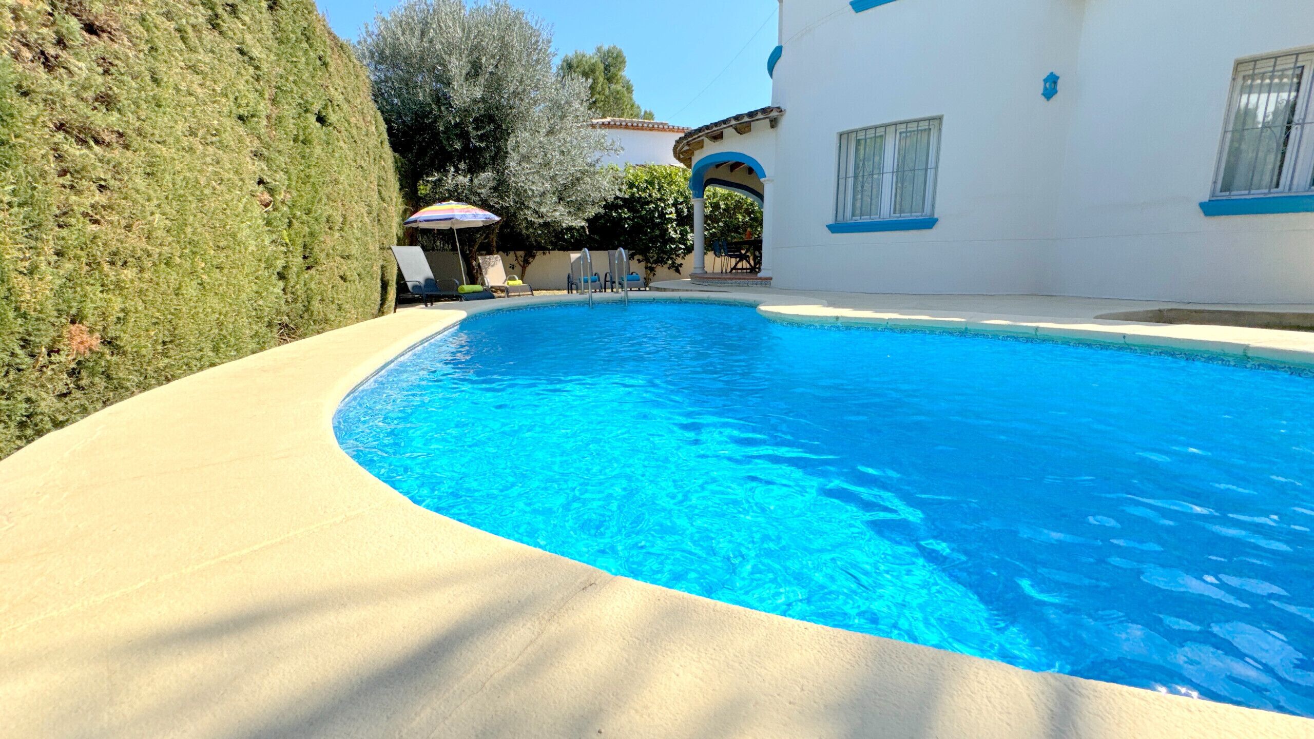Uma piscina com borda curva, rodeada por um deck de cor clara. É possível ver espreguiçadeiras e um guarda-sol ao fundo.