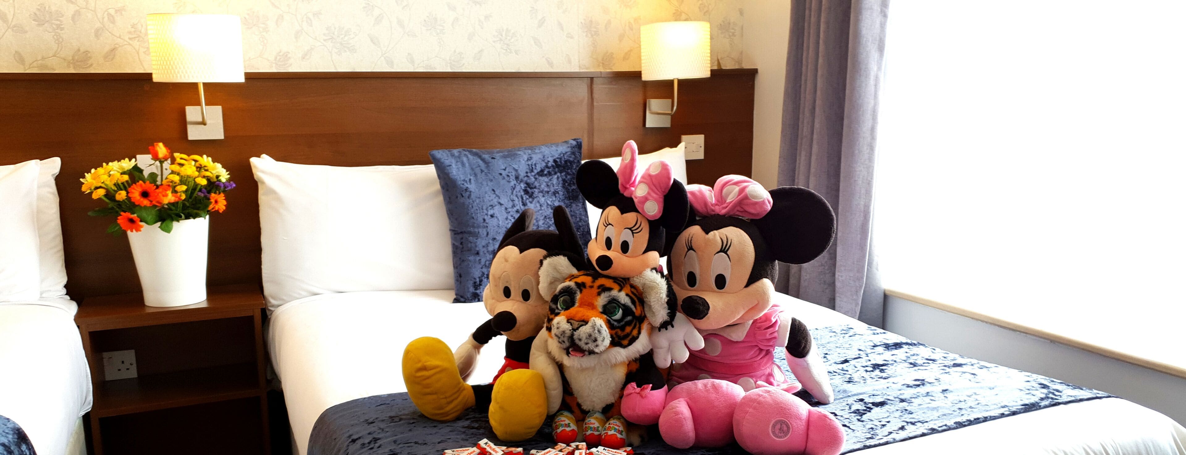 A hotel room with a bed, a nightstand with a lamp, a flower vase, and plush toys on the bed.