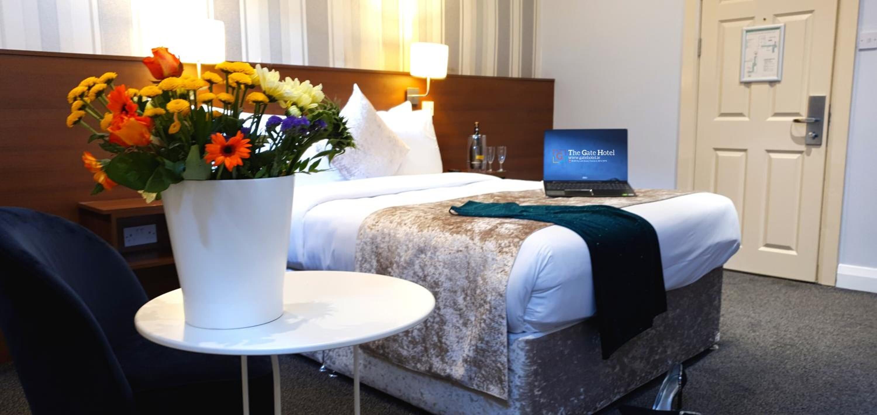 Hotelkamer met een bed, een laptop op het bed, een bloemenvaas en een kleine tafel met een stoel.