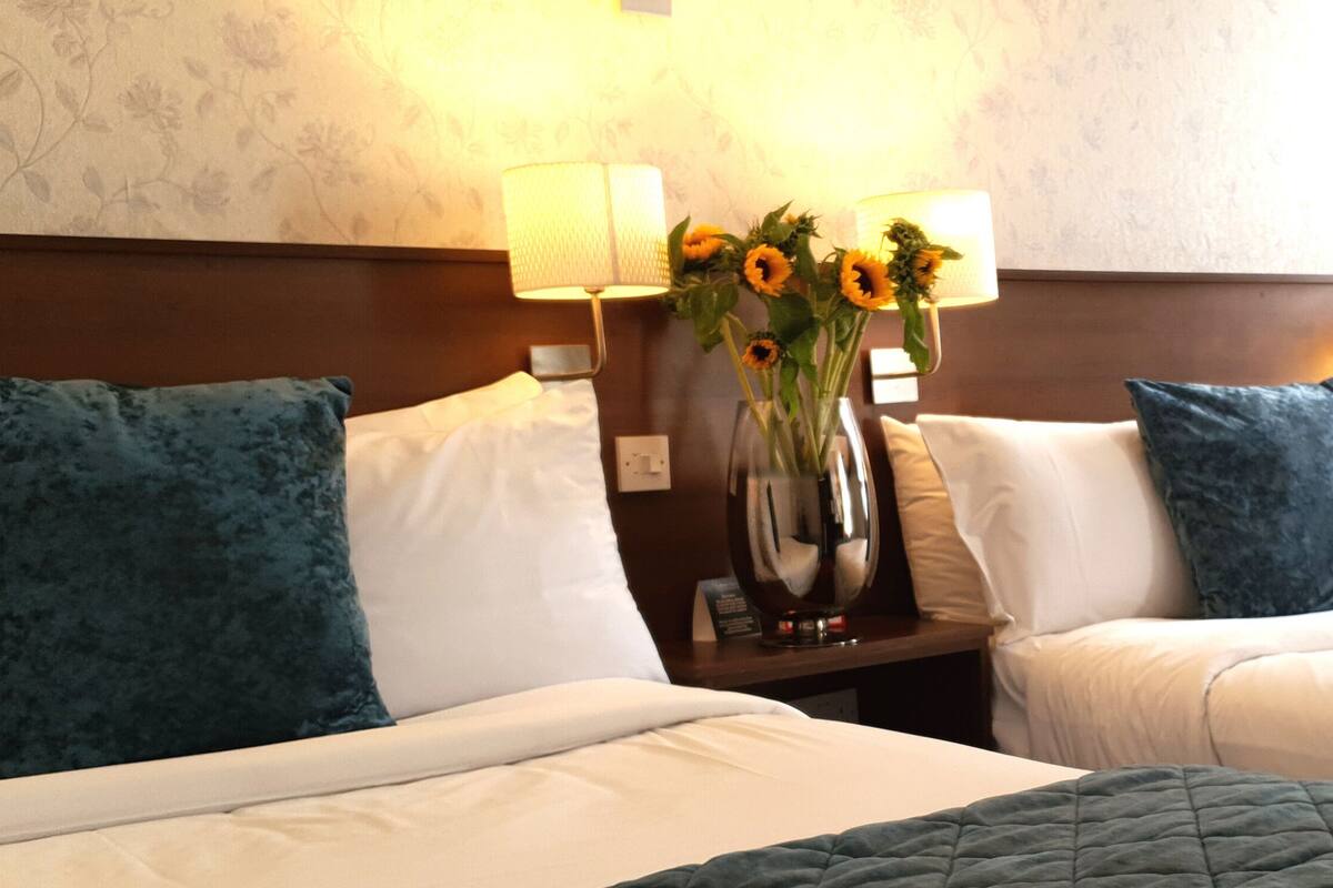 Habitación de hotel con dos camas, cada una con cubrecama azul y blanco, una mesita de noche con un jarrón de girasoles y una lámpara de pared.