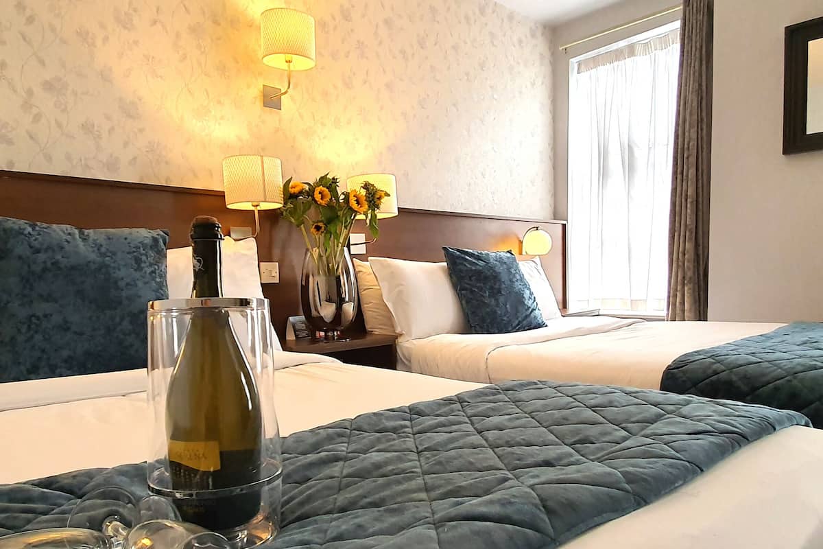 Habitación de hotel con dos camas, cada una con una colcha azul acolchada, almohadas blancas y una jarra con girasoles en la mesita de noche. Hay lámparas fijadas a la pared y un cartel enmarcado.