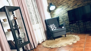 A living room with a dark armchair, a glass display cabinet, a woven rug, and a stone wall. - La Barraca de Culón (Cañada Juncosa)
