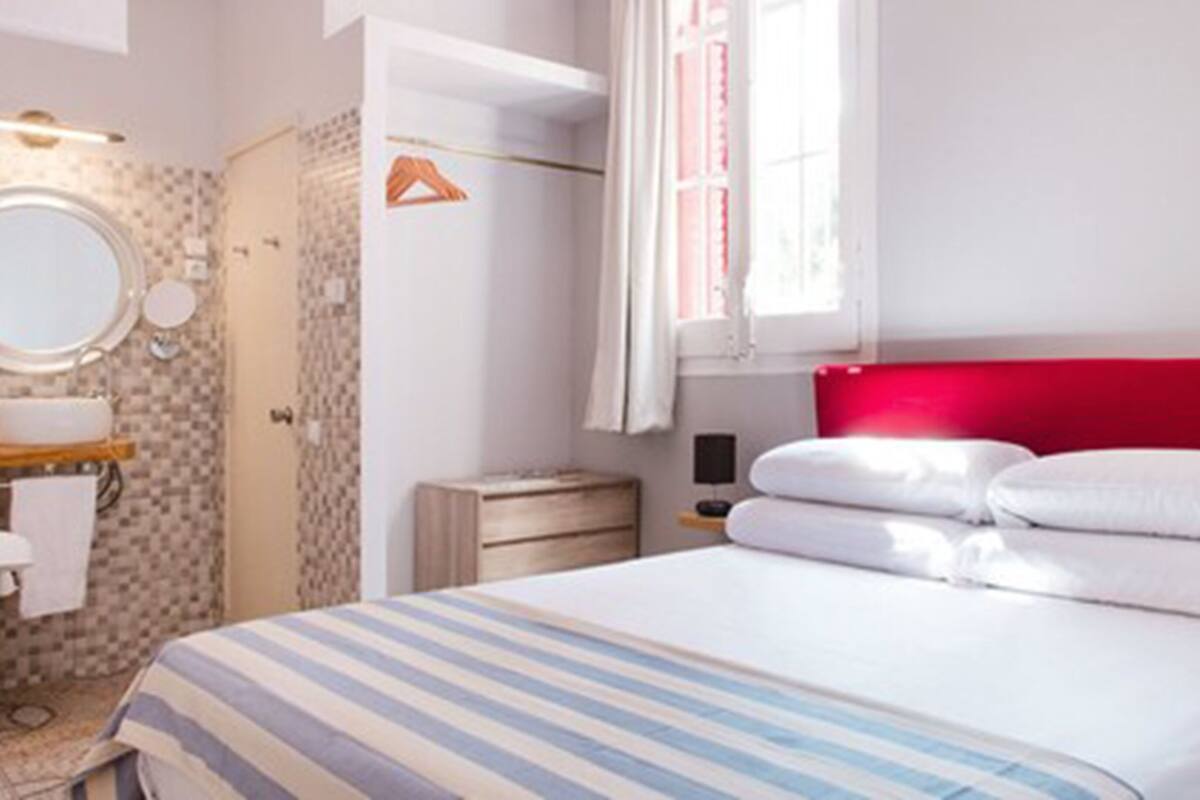 A hotel room with a bed, a bedside table, a mirror, a window with curtains, and a bathroom with a shower and toilet.