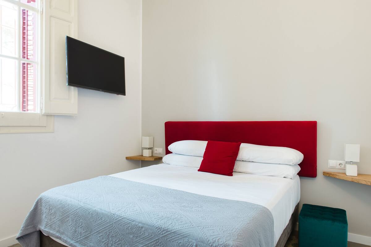 A neatly made bed with a red headboard, a light blue patterned bedspread, and white pillows. There is a flat-screen TV mounted on the wall to the left of the bed.