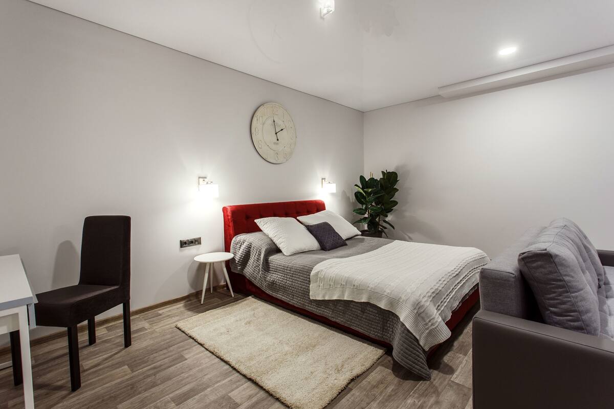 A modern bedroom with a red headboard, a round wall clock, a bedside table, a chair, a grey sofa, and a large plant.