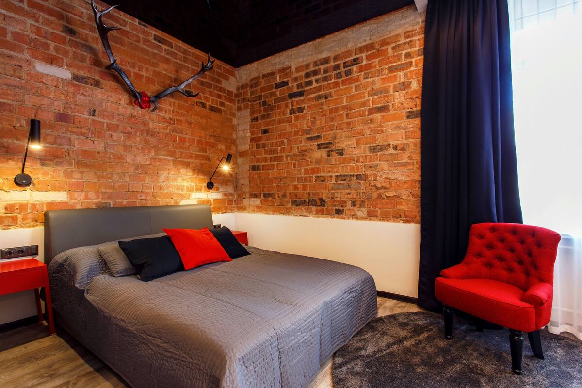 A bedroom with a brick wall, a bed with a grey and red bedspread, a red armchair, and a brick fireplace.