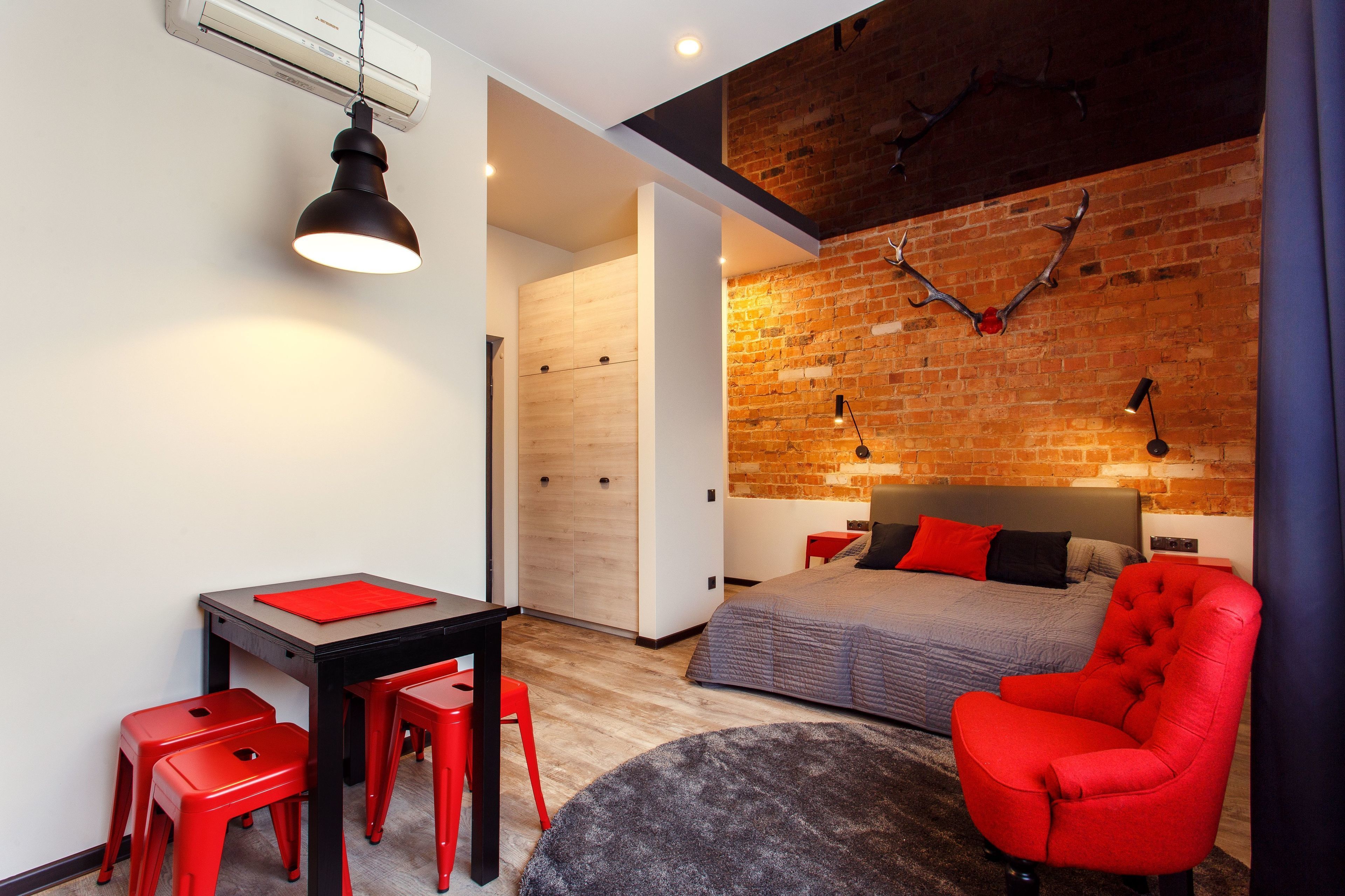A modern bedroom with a brick wall, a bed, a red armchair, a desk with a red chair, and a round rug.