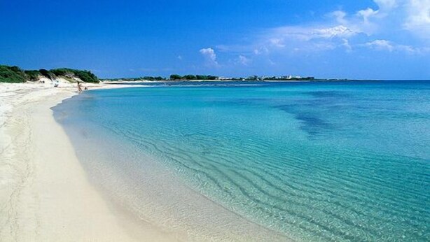 Una playa con agua turquesa cristalina, arena blanca y algunos edificios a lo lejos.