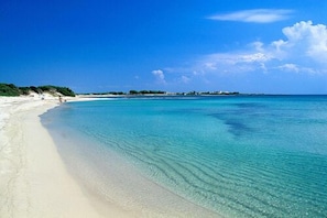 A beach with clear turquoise water, white sand, and a few buildings in the distance.