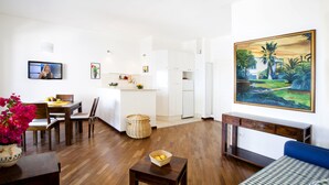 Una sala de estar moderna con sofá, una mesa de centro y un comedor con sillas y una mesa. Hay una cocina con armarios y un refrigerador. Un cuadro adorna la pared, y un televisor está montado en ella.