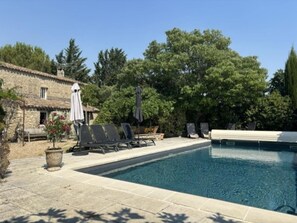 Una piscina con sillones de descanso y sombrillas, rodeada de árboles y un edificio de piedra.