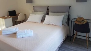 A neatly made bed with white linens, a laptop on a desk, a chair, and a hat on a chair.