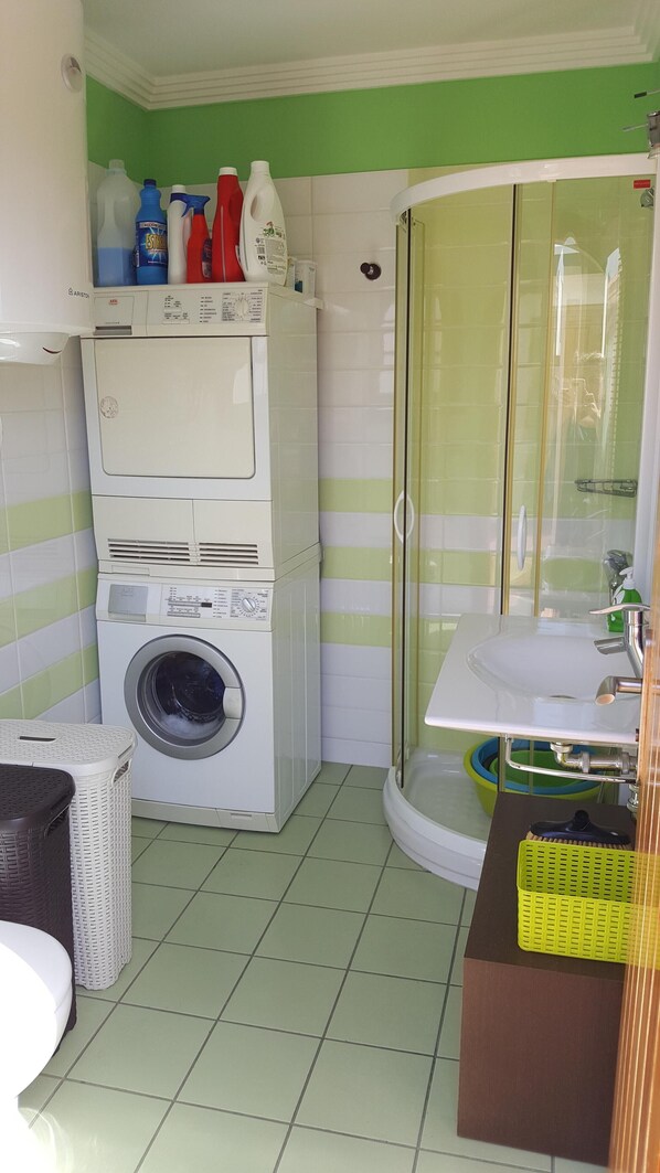 Un lavadero pequeño con lavadora y secadora apiladas, cerramiento de ducha de vidrio y un lavamanos con una cesta verde.