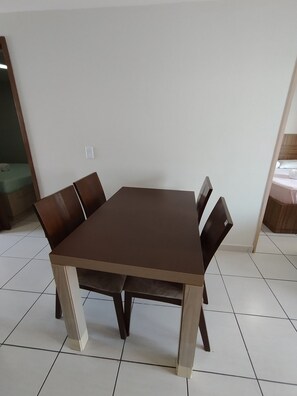 Une table de salle à manger rectangulaire, quatre chaises, un sol carrelé de carreaux blancs et un mur blanc uni.