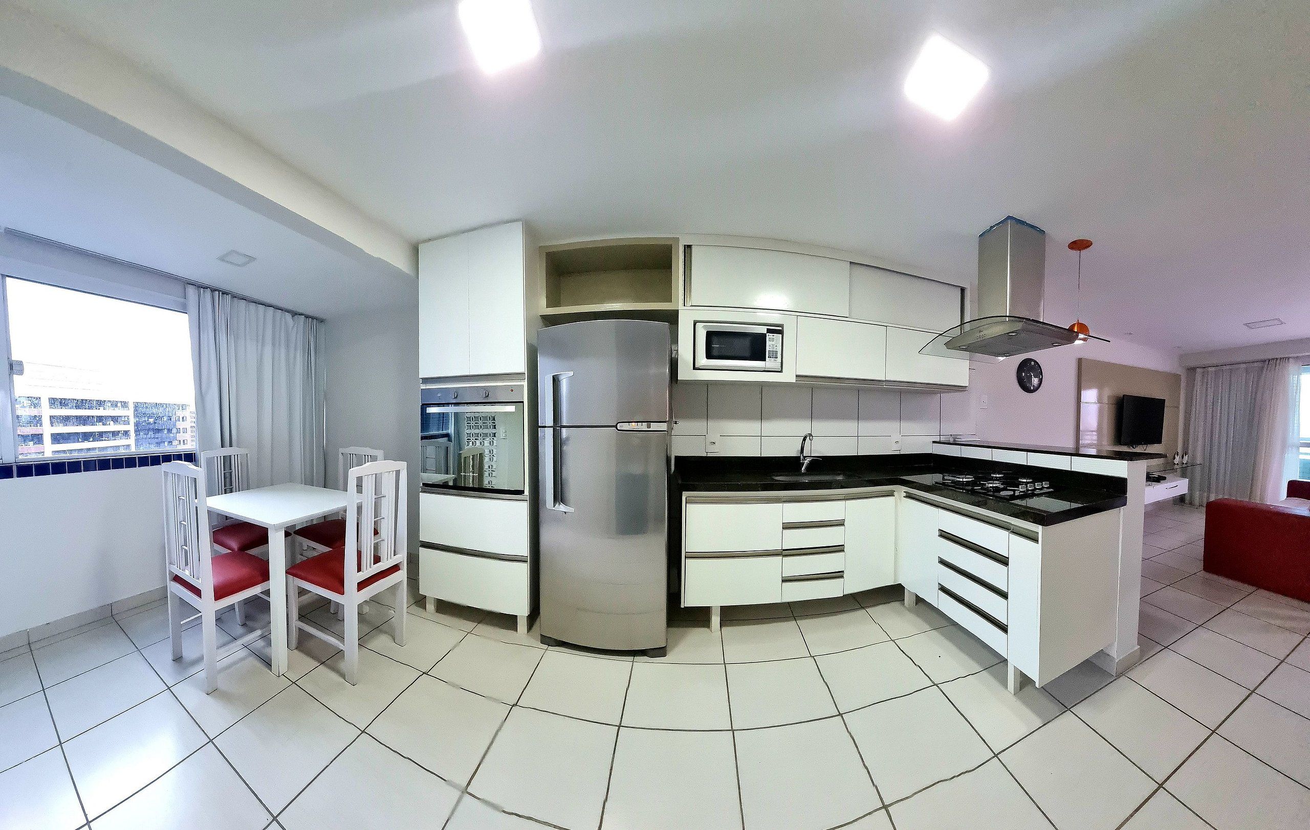 Een moderne keuken met witte kastjes, een roestvrijstalen koelkast, een magnetron en een gasfornuis. Er is een eettafel met rode stoelen en een raam met gordijnen.