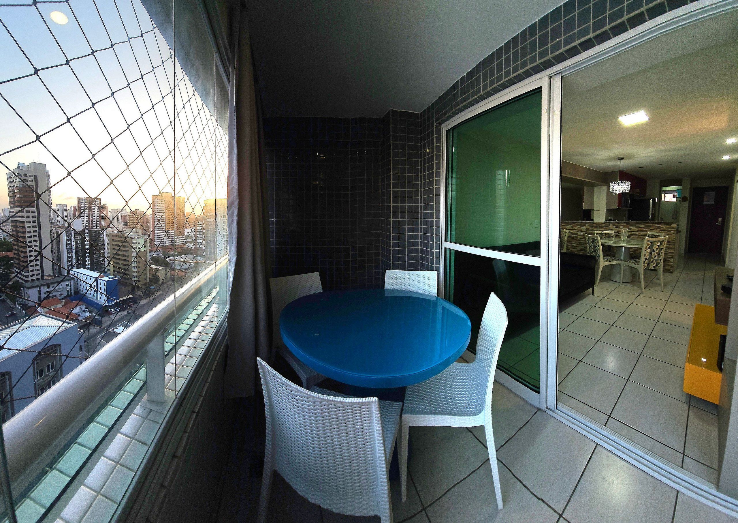 A balcony with a blue table and white chairs, a city view, and a sliding glass door leading to a dining area.