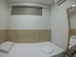 A small, single-bed room with a window, a mirror, and a wall-mounted air conditioning unit.